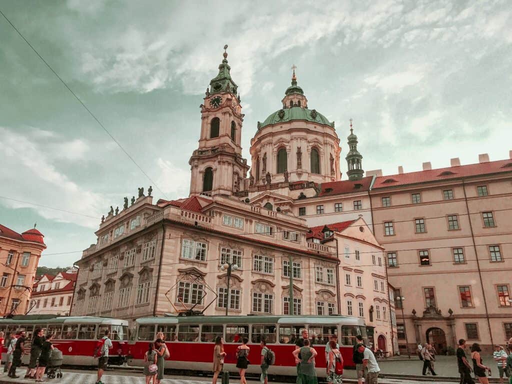 St Nicholas Church in Prague, the building stands with a green dome. In front of it a red and white tram passes, people are standing at the tram stop on the street. 