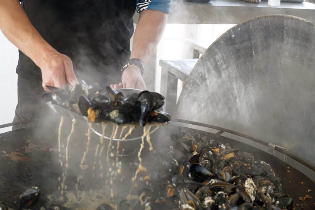 The broth drips from mussels just cooked on a large pan and being transferred to a bowl