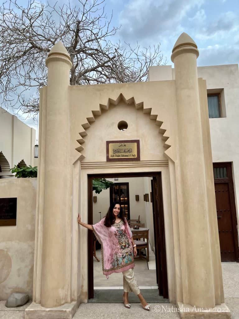 An Indian woman with medium skin and dark shoulder-length hair wearing a pink knee-length kaftan with embroidery and a blue border with off-white pants. Behind her is a sand-colored heritage building in the UAE