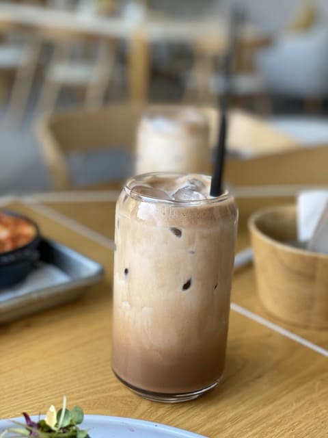 A close up shot of a glass of iced mocha with a straw and two ice cubes in it, placed on a wooden table. 