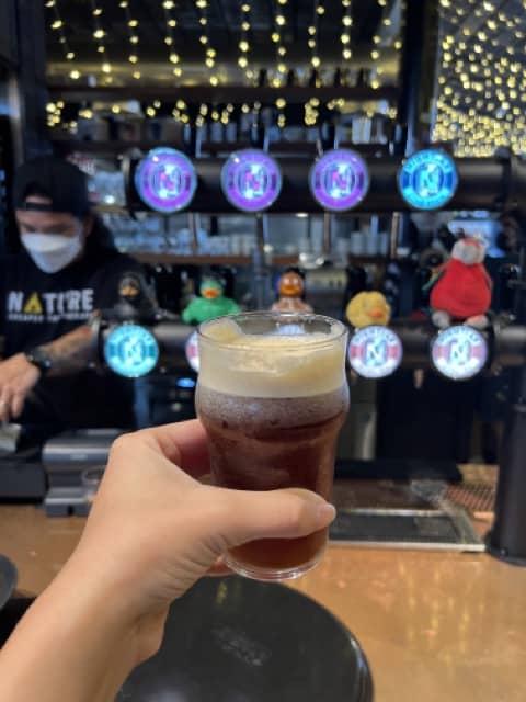A hand holds a glass of cold brew in a cafe, in the backdrop is a cafe counter with cold brew on tap