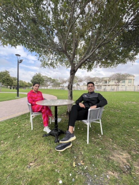 Two people sitting at a table in a park. The man wears a black and gray tracksuit. the woman wears a bright pink tracksuit and her dark hair is in a low bun. They are both Indian and light-skinned. 