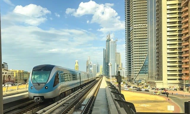 A shiny blue metro runs along a track next to steel gray skyscrapers. Above is a blue sky with clouds.