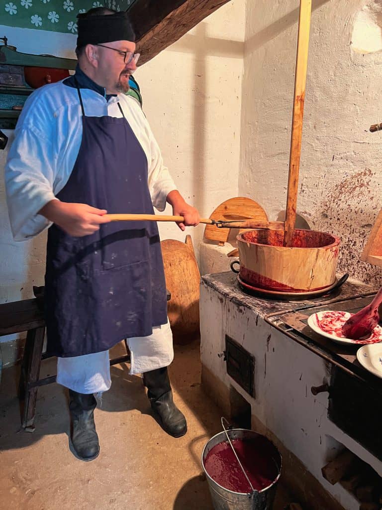 A man in a white shirt and white pants and a navy bue apron and matching headband is making plum jam in a wooden bucket using a wooden paddle. At his feet is a bucket of plum jam. He is wearing glasses. 