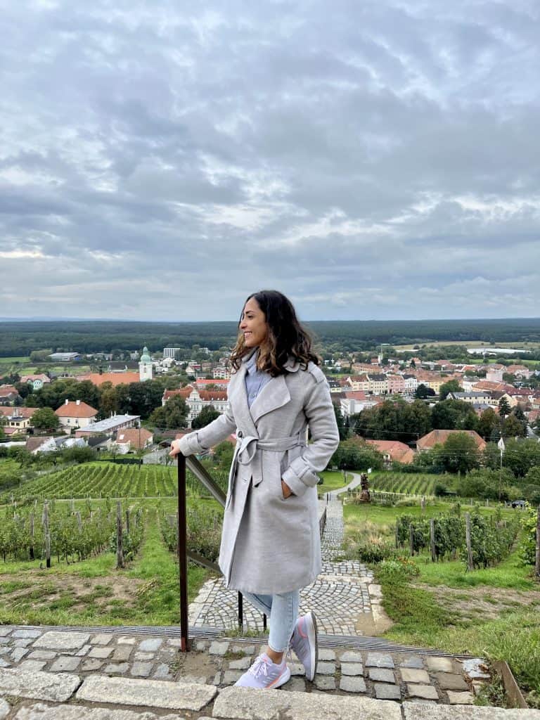 A woman in a light gray trench coat and light blue jeans stands with her right hand in a pocket, her head turned to the side, against the backdrop of a view from a hillside over a town with white buildings and orange red roofs. The hillside is covered with vineyards. She has shoulder length hair and is smiling. 