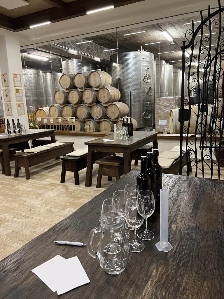 There are 5 wine glasses, a measure, three bottles of wine, and a jar of water placed on a wooden table. This is a tasting room in a winery. In the background are other tables and wine barrels. 