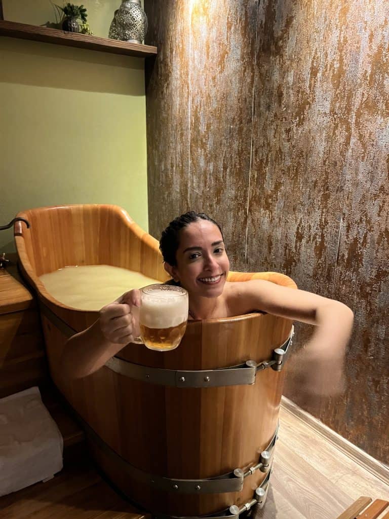 A woman with her hair in a bun is in a wooden tub filled with hops-infused water. She is smiling and has a glass of beer in one hand