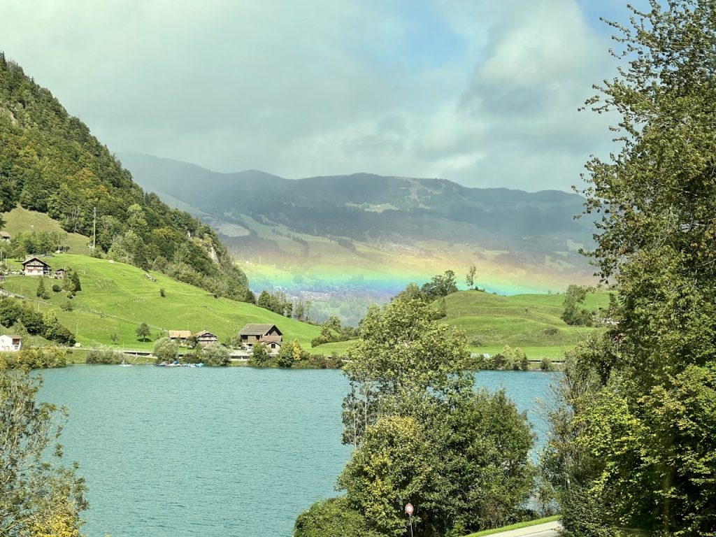 A train window view of rolling green hills beside a blue lake, and a rainbow arching between them