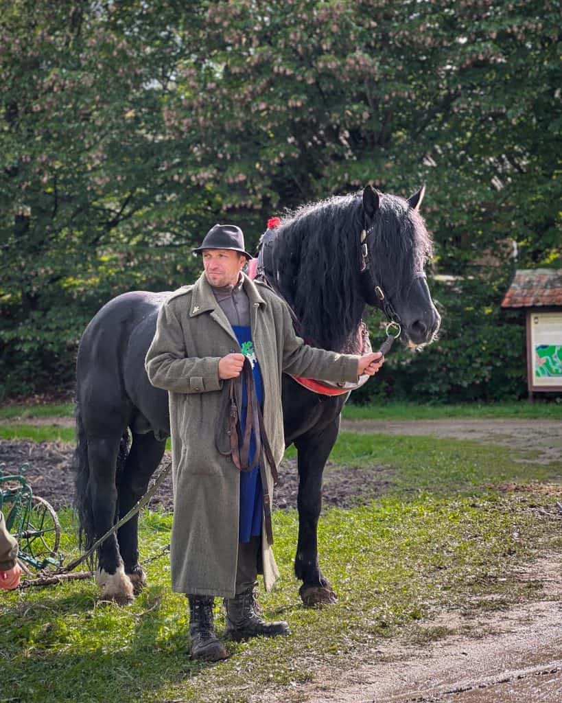 A man wearing a long olive green jacket and a black hat stands next to a black horse, with one hand on the rope attached to a ring secured to the horse's face. The man is Caucasian and wears muddy boots. They are standing outside on the grass and there are trees behind them. 
