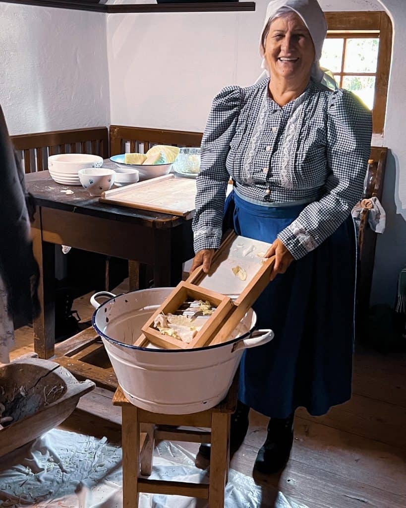 An elderly woman in a blue and white checked dress and white headscarf smiles at the camera while holding a wooden grater in her hand with which she is crushing sauerkraut into a white bucket placed on a wooden stool in front of her. 