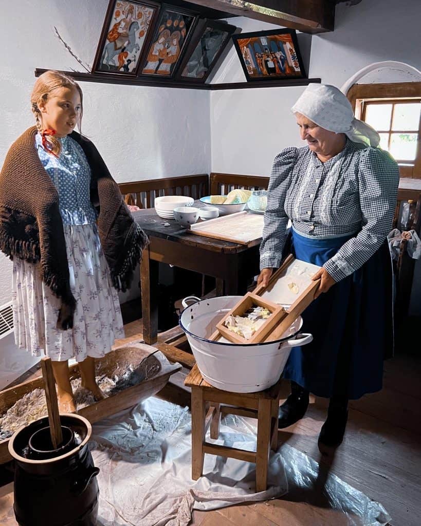 A young girl with light brown hair in two braids stands wearing a blue and white dress and has a brown shawl around her shoulders. She is barefoot and stands in a small wooden tub filled with sauerkraut. Next to her is an older woman wearing a blue and white checked dress and a white headscarf that covers her hair. She is using a wooden crusher ti crush sauerkraut into a white bucket placed on a wooden stool in front of her. They are standing in a small room in a village home with a wooden table in the background on which are bowls and plates. On the wall are medieval style paintings. 