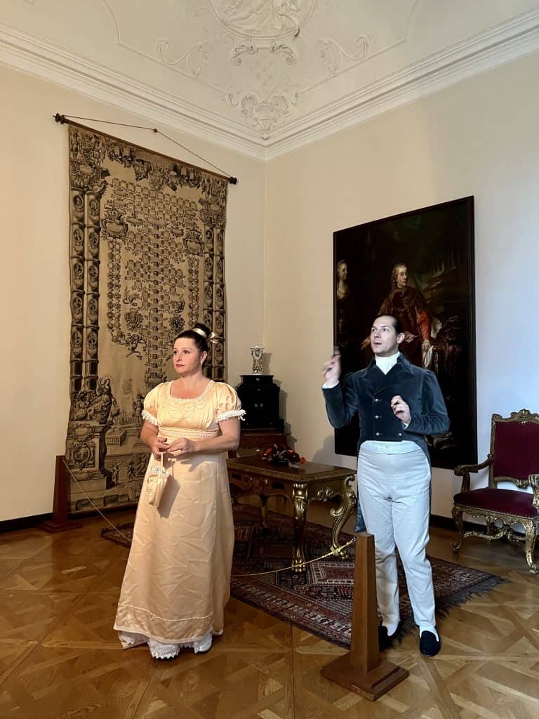 A man dressed in a black coat with tails and cream trousers, his hair pulled back in a ponytail, stands with a woman in a peach satin gown, her hair in a high bun. They are in one of the rooms in the chateau and are pointing at paintings and exhibits and talking about them. 