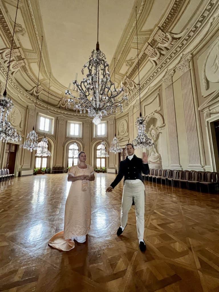A man dressed in a black coat with tails and cream trousers, his hair pulled back in a ponytail, stands with a woman in a peach satin gown, her hair in a high bun. They are looking up at the crystal chandeliers in a grand ballroom of sorts. 
