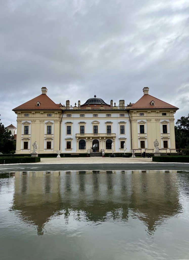 A grand Baroque chateau with a white central building and two adjacent cream colored buildings on either side, with orange-red roofs and a central domes, stands before a pool of water. The chateau is reflected in the pool. The sky above is gray and dramatic. 