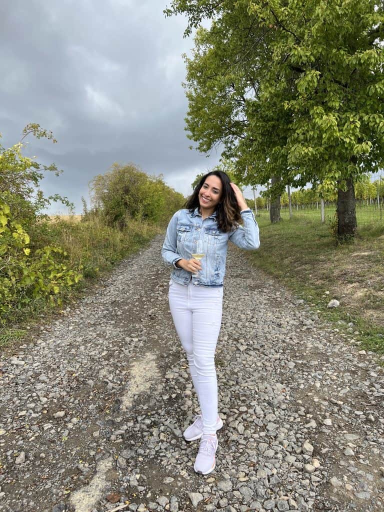 A girl stands with a glass of white wine in her right hand, on a gravel path beside a vineyard in Moravian Tuscany. She has shoulder length black hair, and her left hand is on her hair pushing it away from her face. She is wearing fitted white jeans and a light blue denim jacket. 