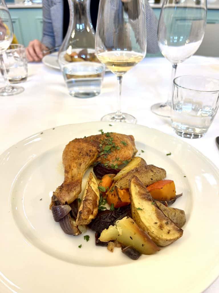 A plate of roasted chicken with a side of roasted carrots and potatoes, next to a glass of white wine