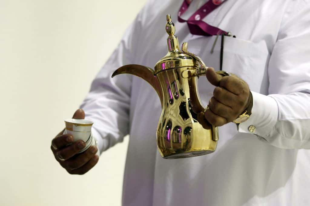 A man pours coffee from a Dallah coffee pot with a long spout into a small cup called finjan