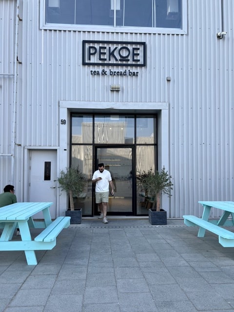 A man in a white t-shirt and cream shorts is looking at his phone as he steps out of a glass door of what appears to be a cafe with the name on top Pekoe Tea and Bread Bar