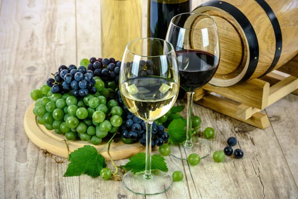 Two glasses of wine, one with white wine, the other with red wine, placed on a wooden surface. Behind them is a bunch of green and another bunch of red grapes.
