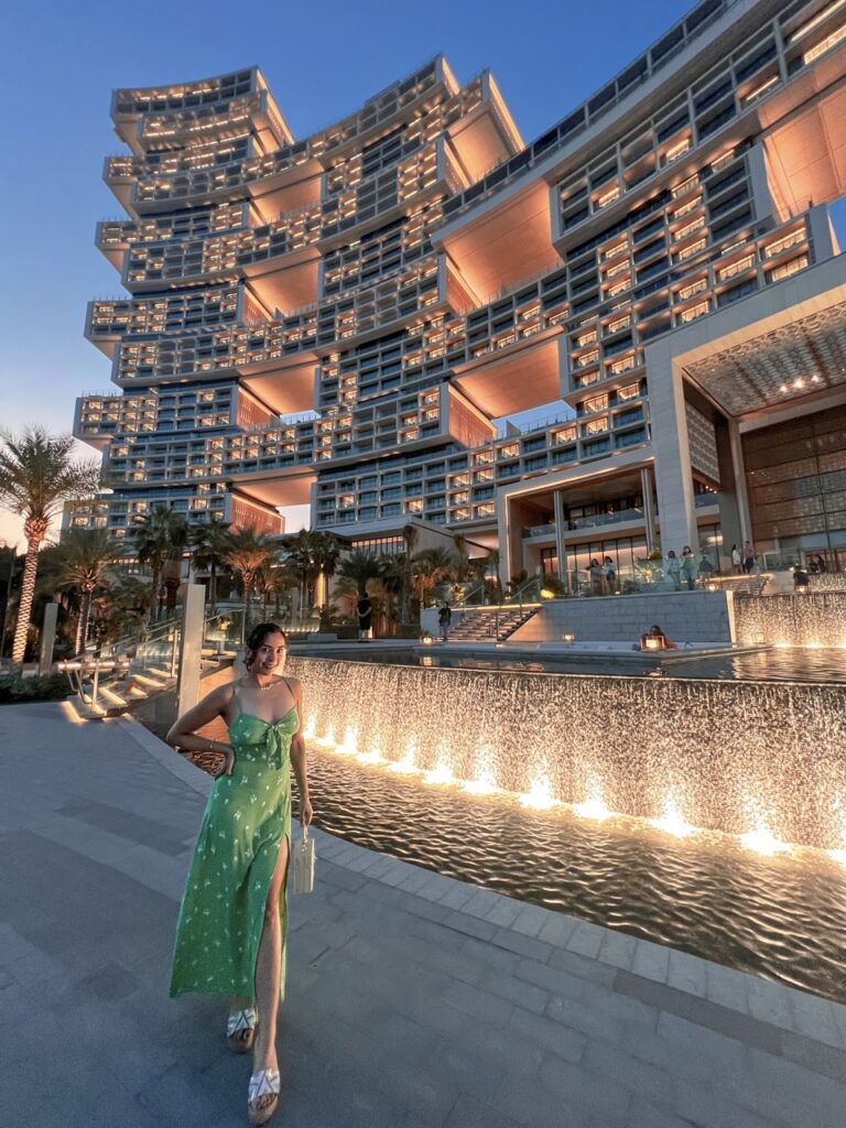 The contemporary building of the Atlantis The Royal lit up with warm lighting. In front of it a girl stands posing with an arm on a hip, wearing a green dress, her hair in a bun.