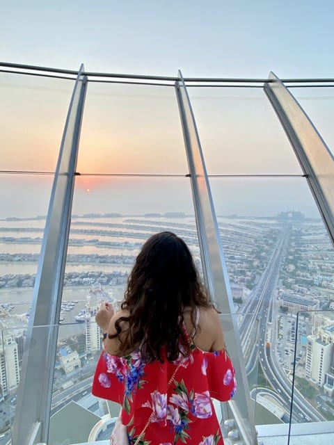 A girl with curly brown hair in a red dress looks out of a panoramic glass window at the outdoor observatory of the Palm Tower. She is looking at a sunset view of the manmade palm-shaped islands of Palm Jumeirah in Dubai 
