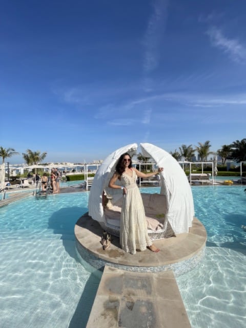 A girl in a cream-colored crochet maxi poses with one hand against a pod-shaped cabana on a terraced pool. A walkway leads to the cabana. She has dark hair, is light-skinned, and is smiling. The sky behind her is a bright blue. 