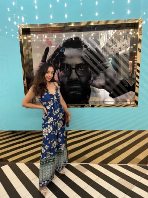 A girl posing in front of a large photograph of Italian chef Massimo Bottura on a blue wall at the entrance of the restaurant Torno Subito in Dubai. She has dark hair which is down and wears a long blue silk jumpsuit with a white floral design. Her right hand is on her hip. 