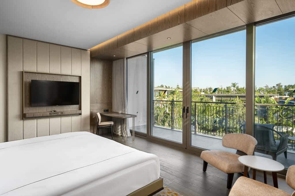 A bedroom with a TV on the wall and view of pool and trees through floor-to-ceiling glass doors. 