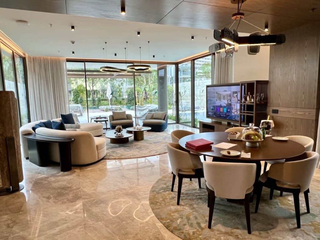 A living room that begins with a dining area with a round dining table and six chairs, followed by a seating area with a couch, two chairs, and nested coffee tables. All around are floor-to-ceiling glass windows that look upon a pool terrace. 