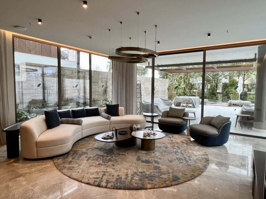 A living room that begins with a dining area with a round dining table and six chairs, followed by a seating area with a couch, two chairs, and nested coffee tables. All around are floor-to-ceiling glass windows that look upon a pool terrace. 