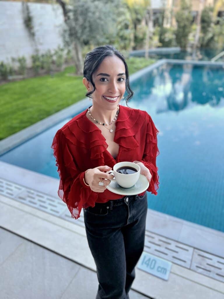 A woman in a red blouse and black jeans holding a cup of black coffee. behind her is a pool and trees.  