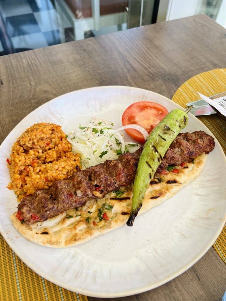 A plate of a kebab with a side of bulgur, some white onions and a grilled tomato, all topped with a long green chilli. 