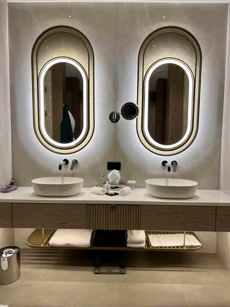 A bathroom close-uo of twin vanities with backlit oblong mirror and two sinks. It's all marble and ivory-colored. 